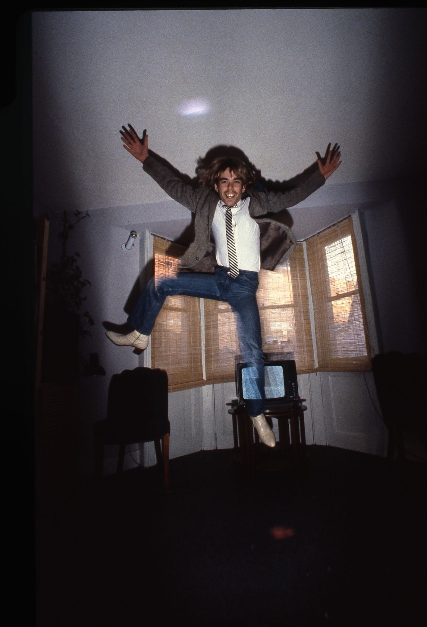A smiling man in a jacket and jeans leaps in front of a window.