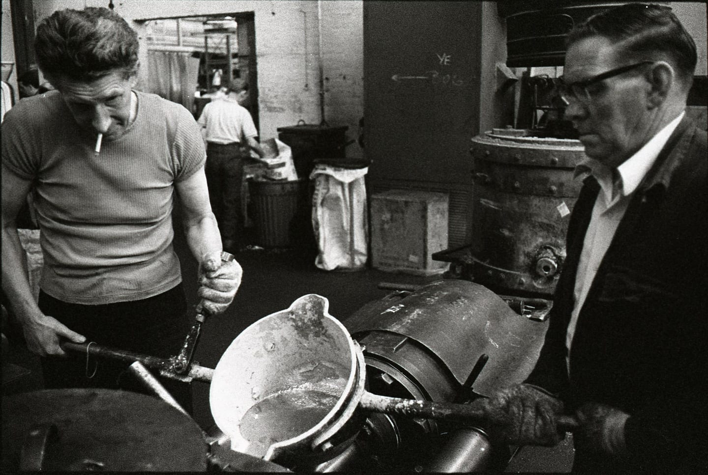 Two workers handle a ladle beside industrial machinery in a black-and-white workshop photograph.