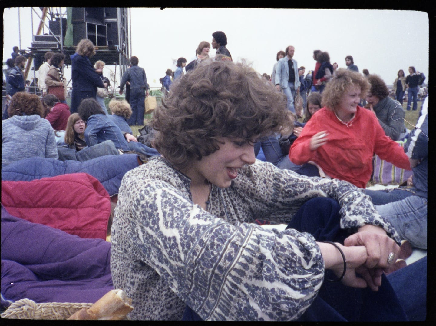 A young man in a patterned shirt sits among blankets and a crowd near stage equipment.