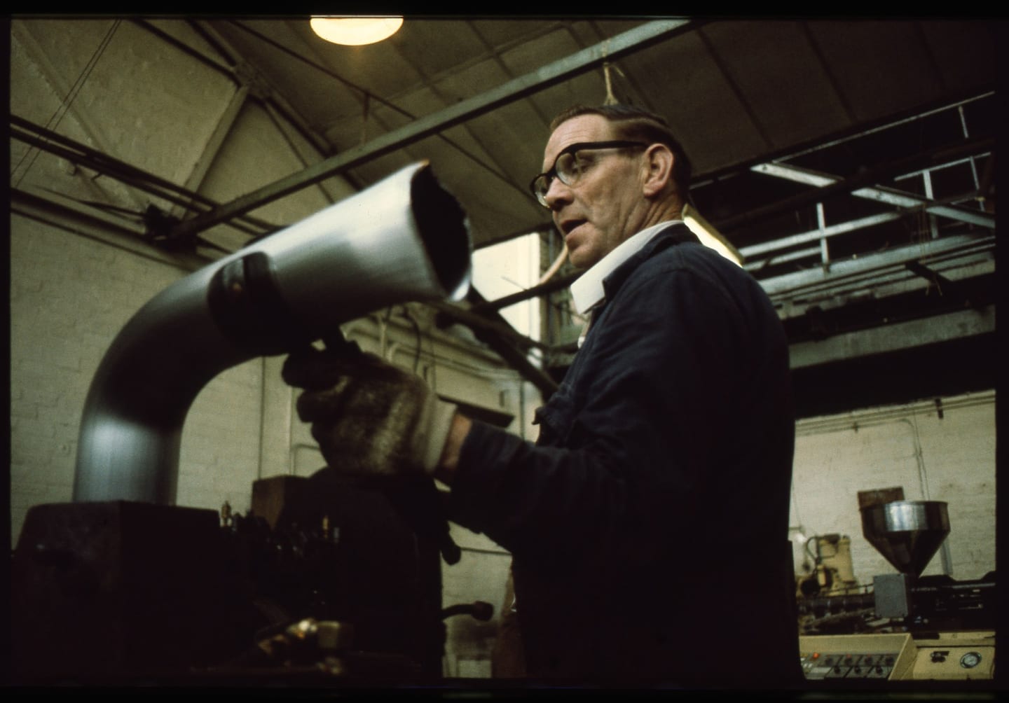 A bespectacled worker wearing protective gloves holds a curved pipe inside a workshop.