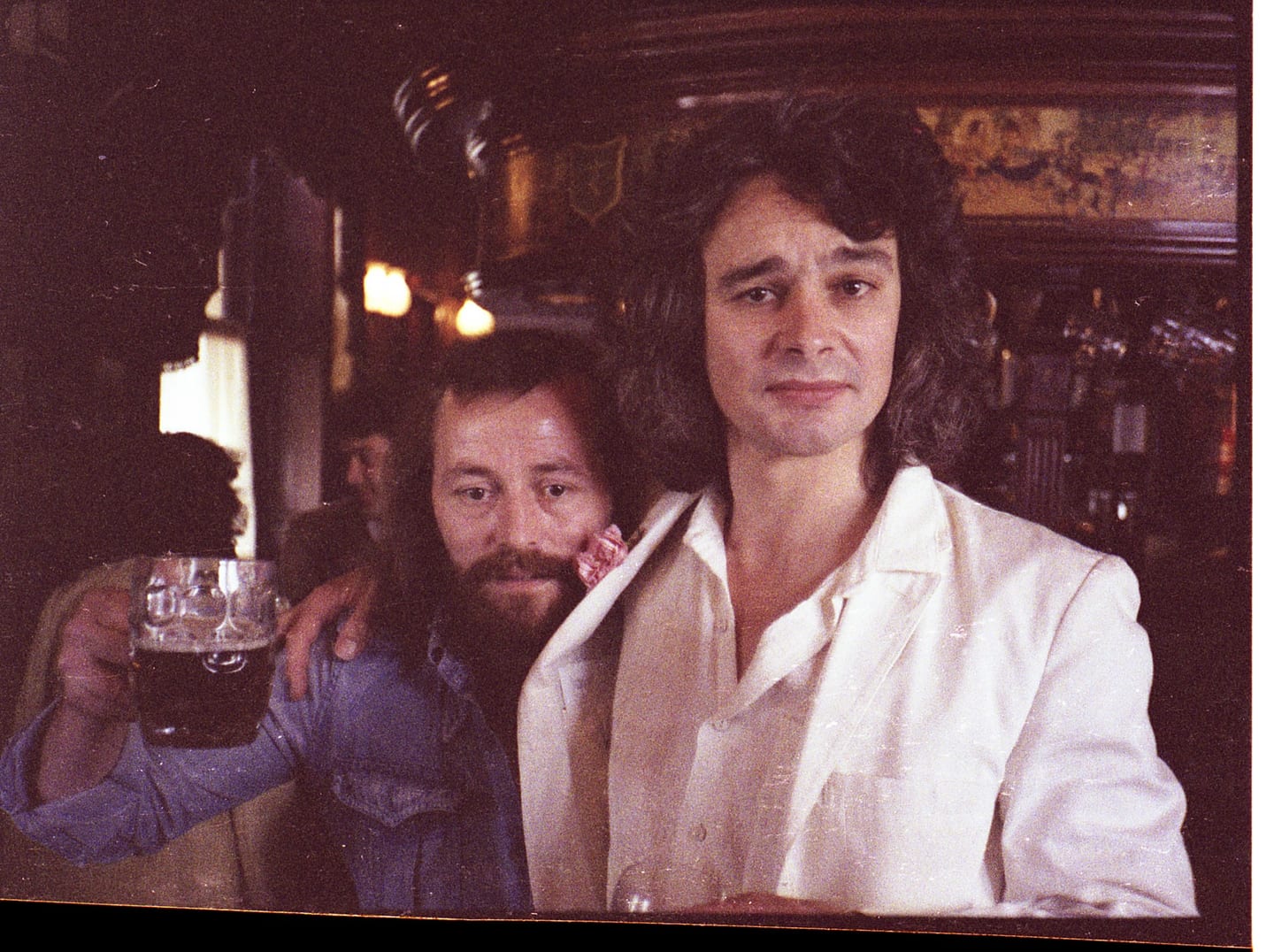 Two men stand together in a pub, one holding a glass of beer.