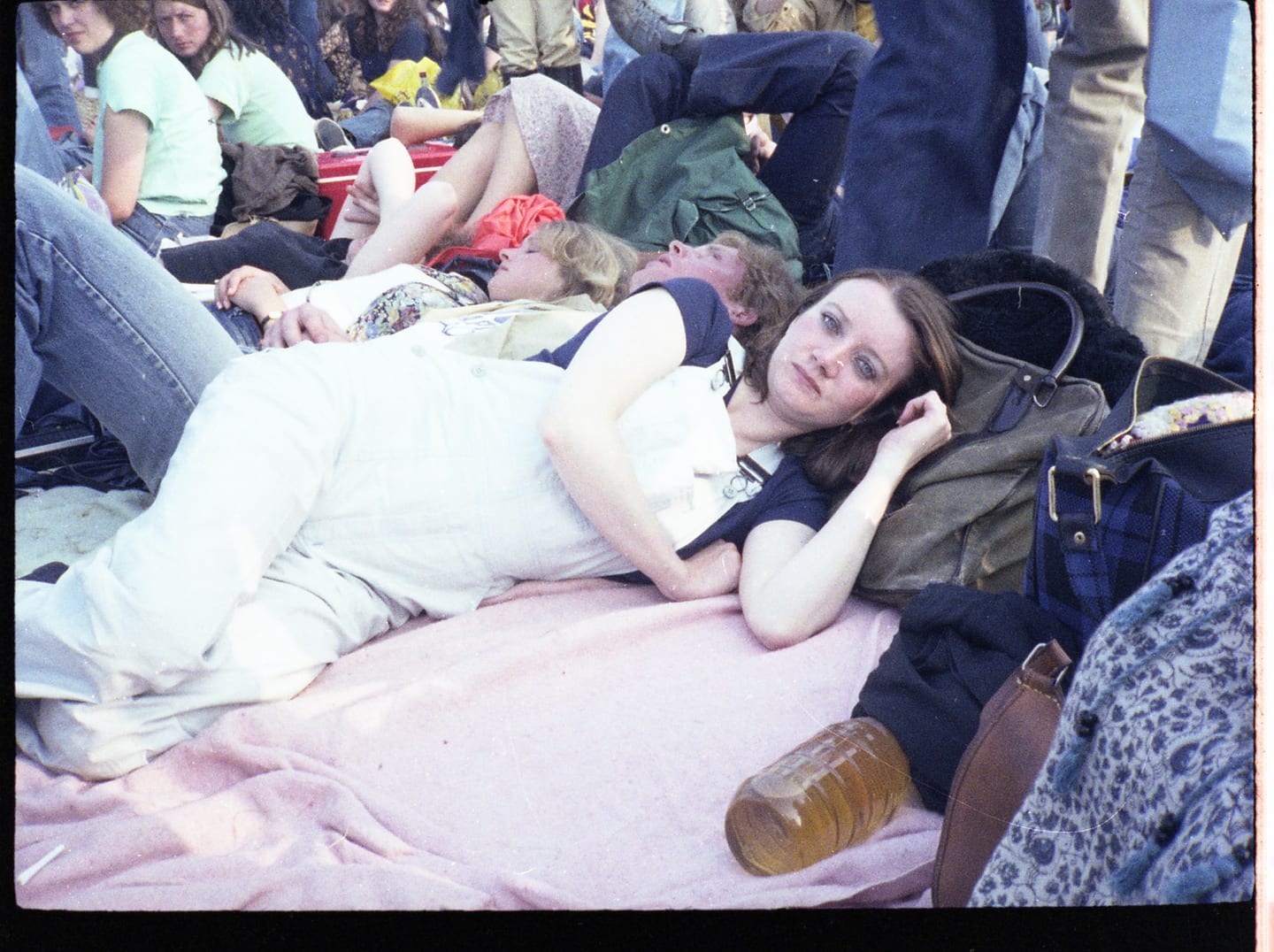 A woman reclines on a pink blanket among bags and people at an outdoor gathering.