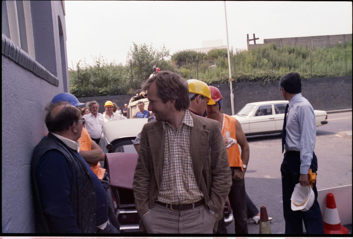 A man in a checked jacket stands among a group near parked cars and traffic cones.