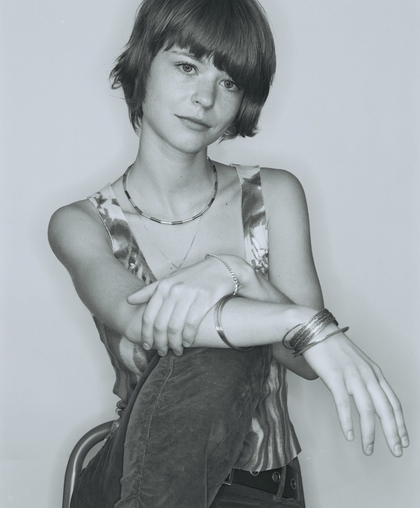 A woman with short hair rests her arms across a raised knee, wearing necklaces and bangles.