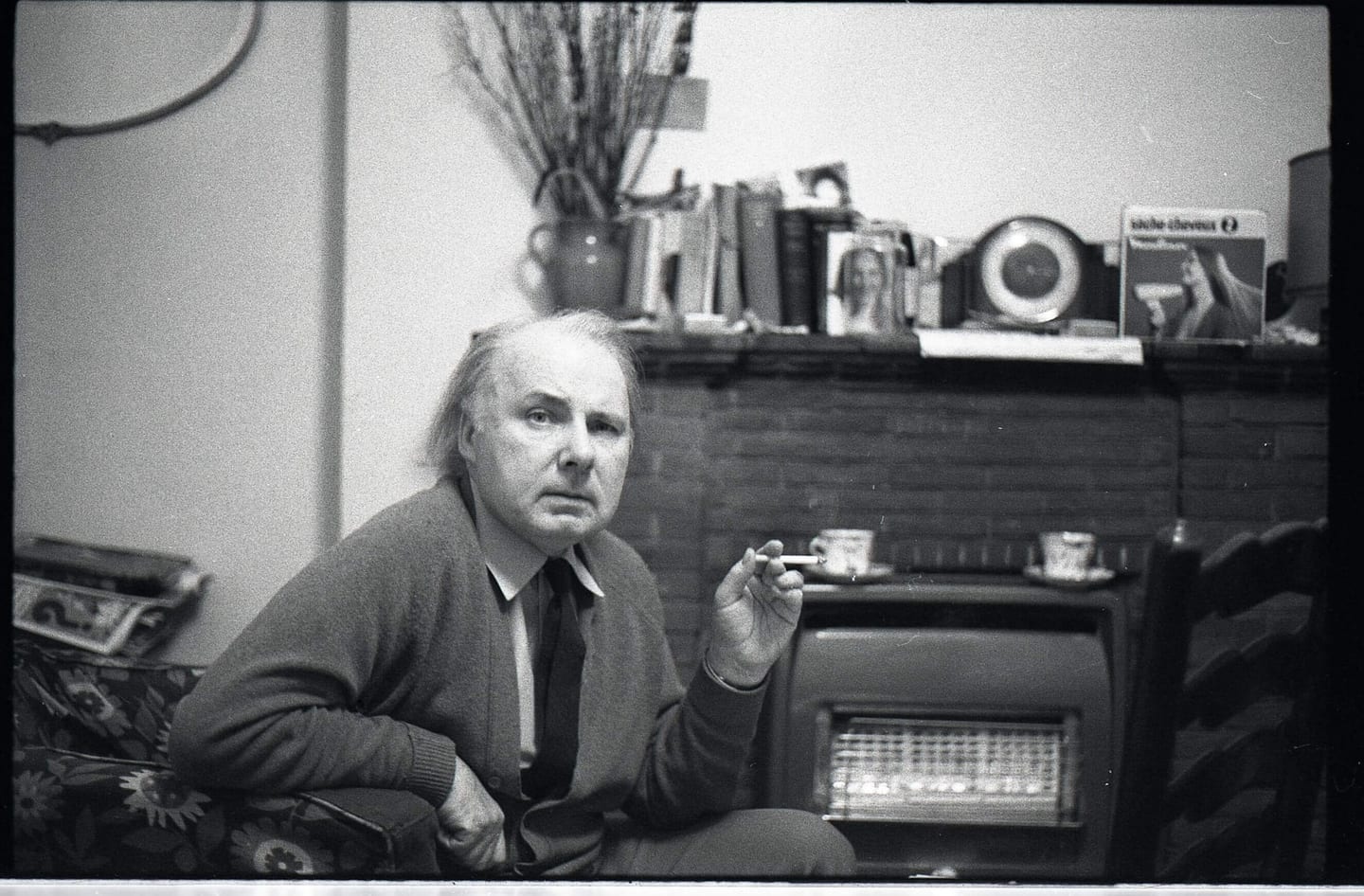 A man sits in an armchair holding a cigarette near a mantelpiece lined with books.