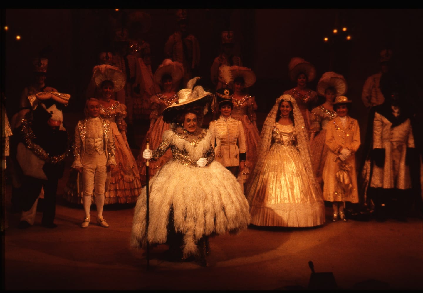 A group of performers in elaborate dresses and hats stand together on a lit stage.