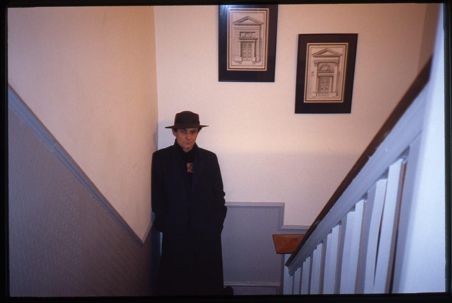 A man in a dark overcoat and hat stands on a staircase beneath framed pictures.