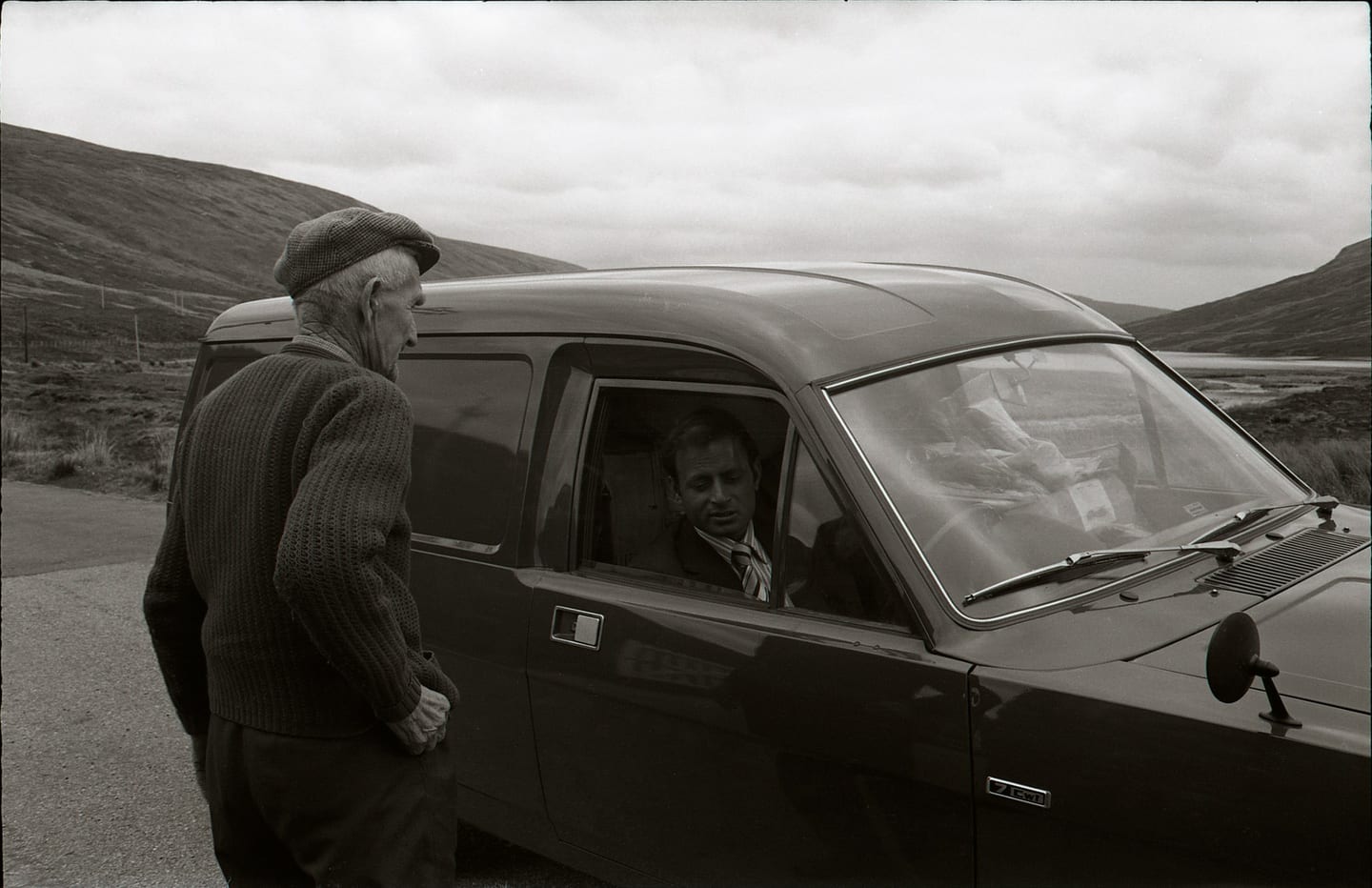 A man in a flat cap stands beside a car speaking to a seated occupant, with hills beyond.